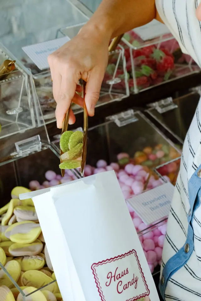 Gold tongs drop green sour gummies into a white Haus Candy bag
