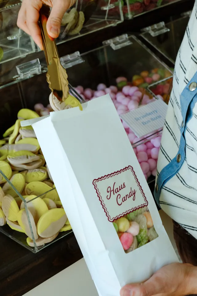 Gold tongs drop candy into a white Haus Candy bag above bins of banana toffee ovals and Strawberry Bon Bons