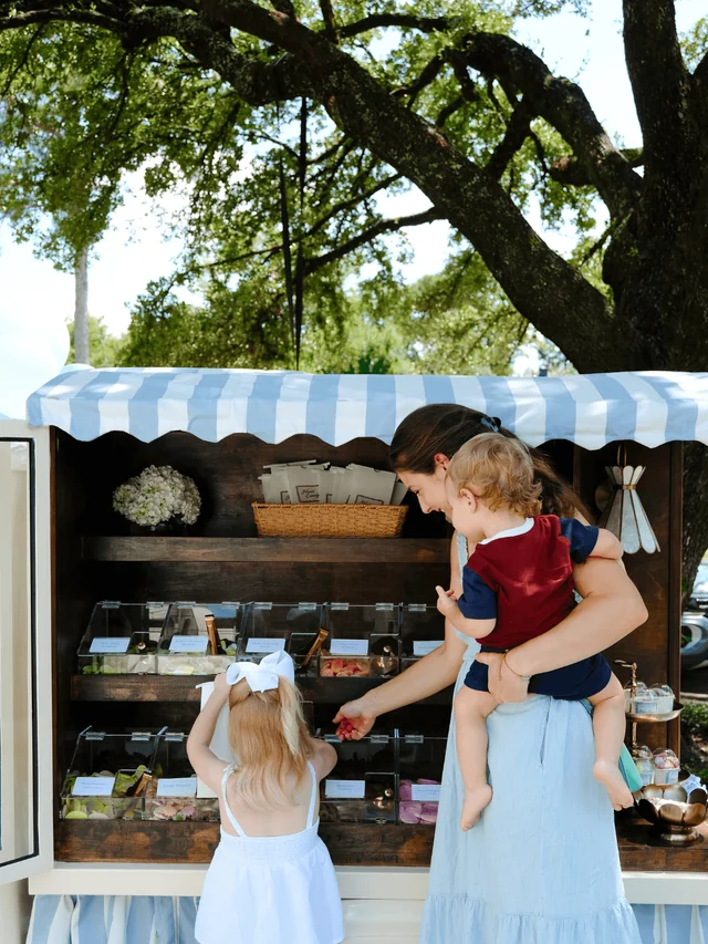 A woman holding a toddler helps a young guest choose candy from the Cart