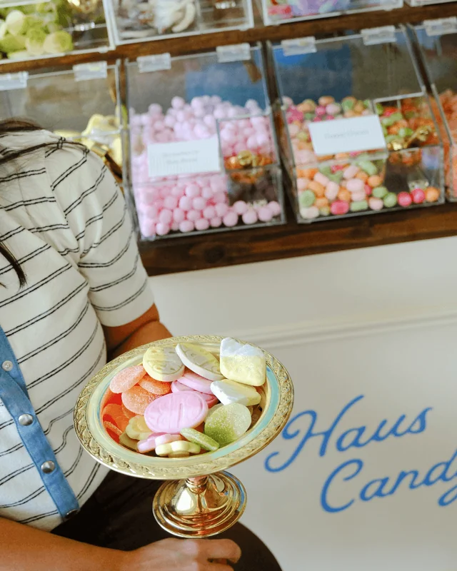 A guest holds a gold pedestal dish of colorful sweets beside the Haus Candy Cart