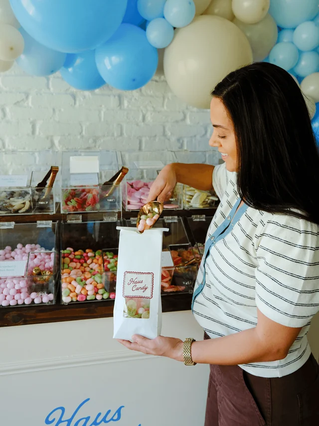 A guest uses a gold scoop to fill a white Haus Candy bag beside clear candy bins